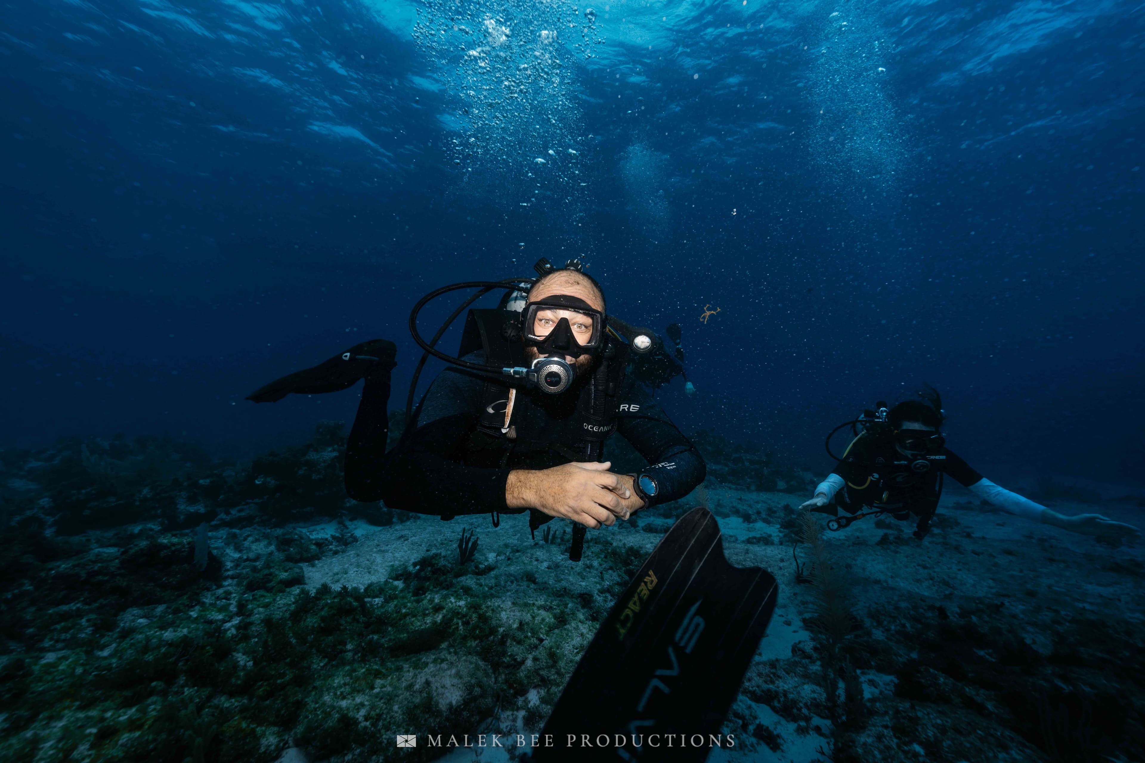 Scuba diver in black gear swims near the seabed with bubbles rising in blue water.