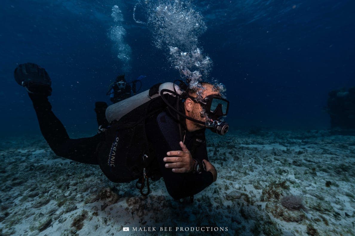 Scuba diver swims through deep blue water, exhaling bubbles above a sandy, rocky seabed.