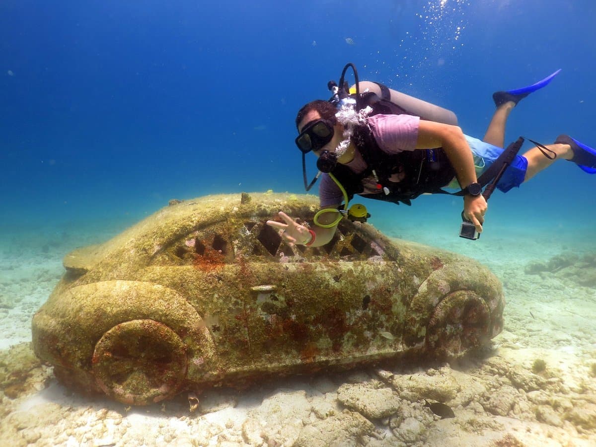 Scuba diver swimming next to a coral-encrusted car sculpture on the sandy ocean floor.